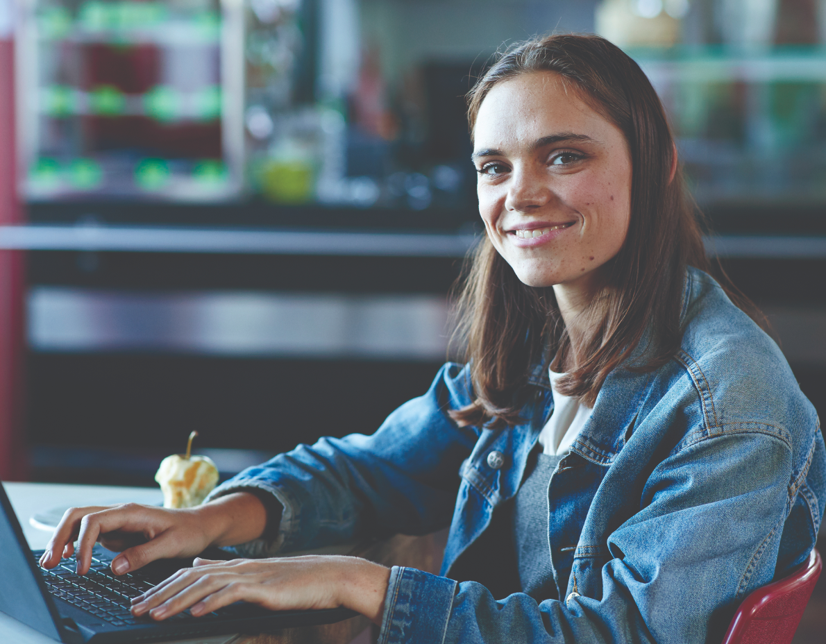 A young student smiles at the camera while working on a laptop.