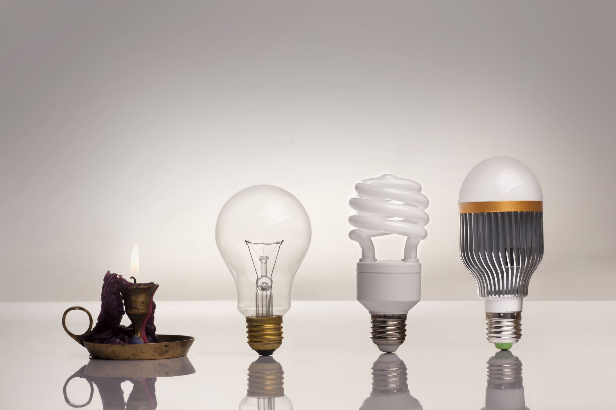 A candle, an incandescent bulb, an LED bulb, and a RGB bulb lined up on a table.