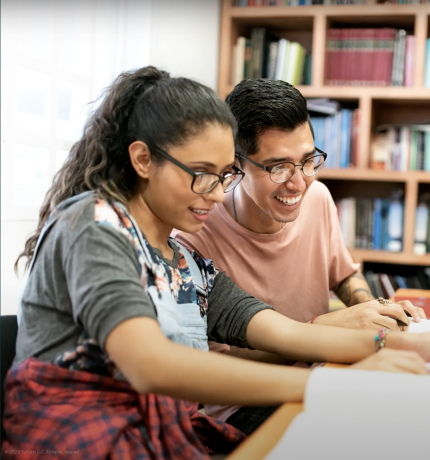 Two students working together on a laptop at a table in the library. 